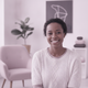 A happy mother in her late 20s, with a warm smile, wearing soft pale mauve and white clothing. The background is softly blurr